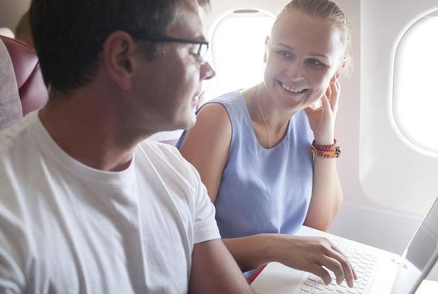 Happy young people traveling by plane.  Woman talking to a man while using laptop. Bright sunlight in airplane windows